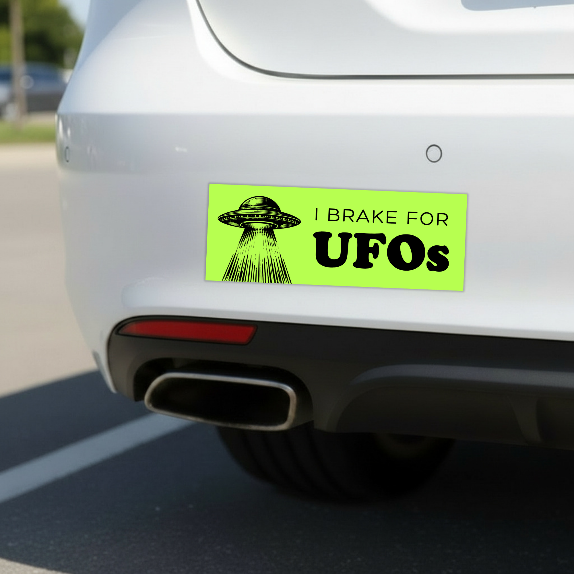 Sleazy Greetings funny bumper magnet displayed on the back of a white car featuring the phrase ‘I Brake For UFOs’ in bold black typography on a neon green background with a vintage-style flying saucer illustration beaming light downward. Humorous novelty car magnet with retro sci-fi aesthetic and alien conspiracy humor shown in real-life automotive use.