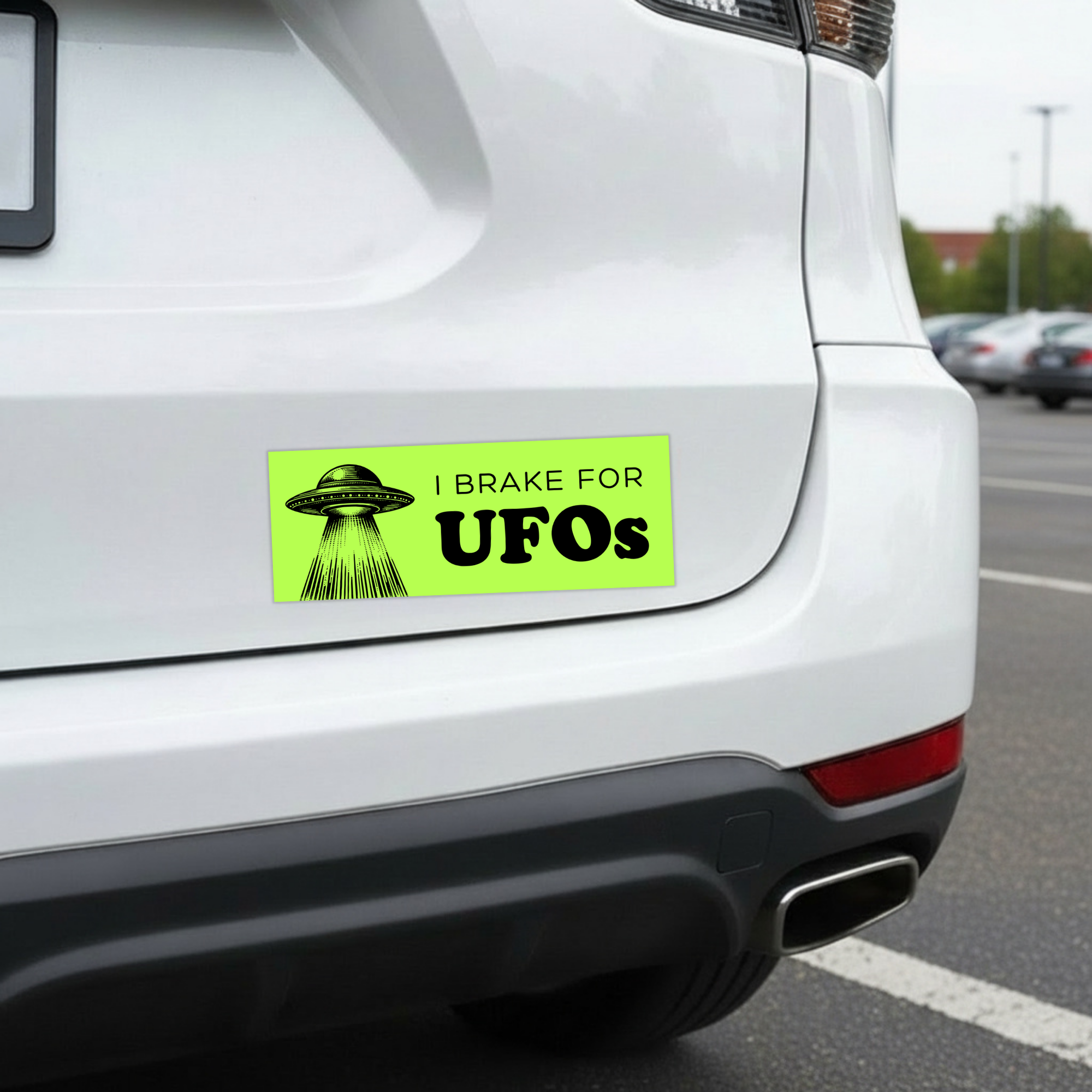 Sleazy Greetings funny bumper magnet displayed on the back of a white car featuring the phrase ‘I Brake For UFOs’ in bold black typography on a neon green background with a vintage-style flying saucer illustration beaming light downward. Humorous novelty car magnet with retro sci-fi aesthetic and alien conspiracy humor shown in real-life automotive use.