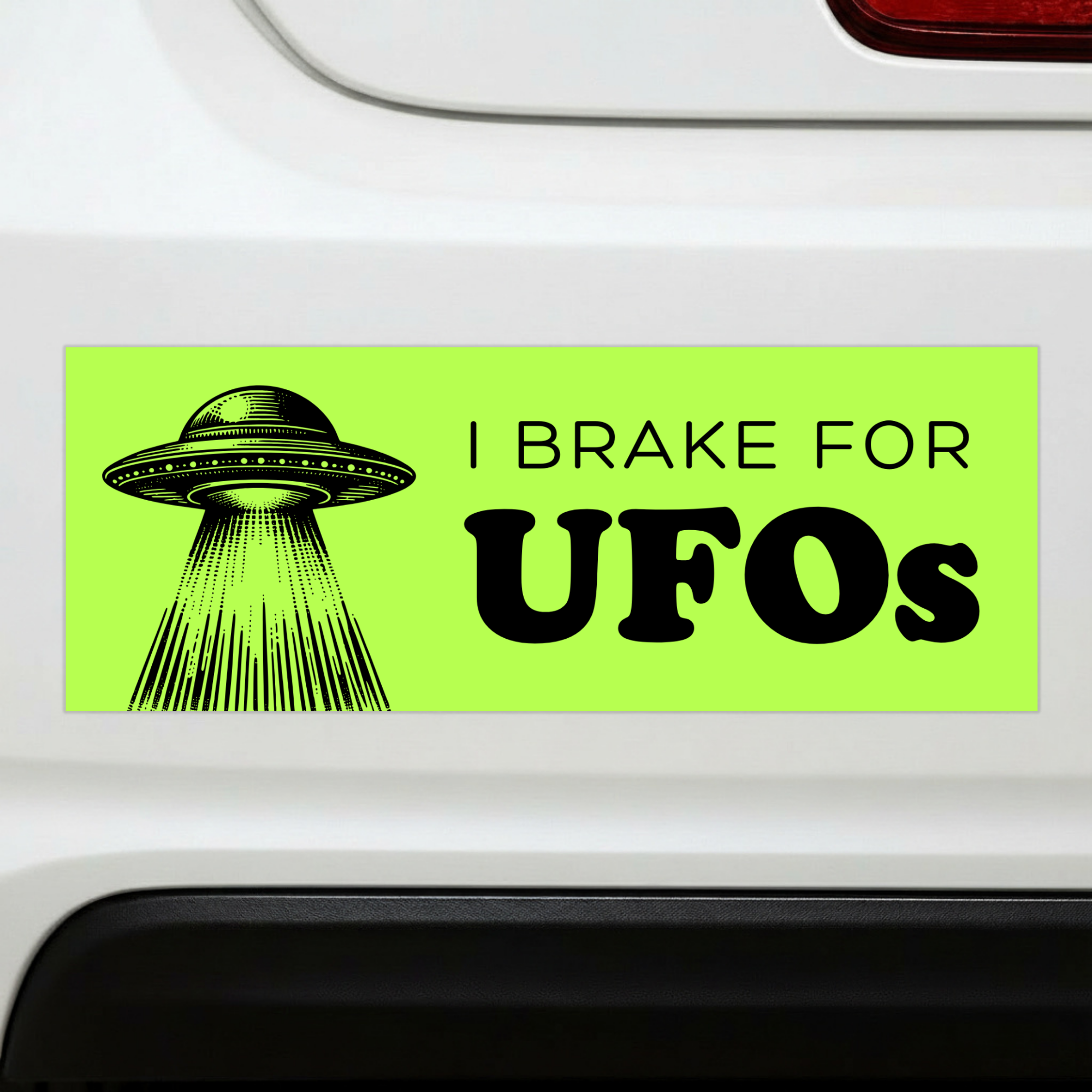 Sleazy Greetings funny bumper magnet displayed on the back of a white car featuring the phrase ‘I Brake For UFOs’ in bold black typography on a neon green background with a vintage-style flying saucer illustration beaming light downward. Humorous novelty car magnet with retro sci-fi aesthetic and alien conspiracy humor shown in real-life automotive use.