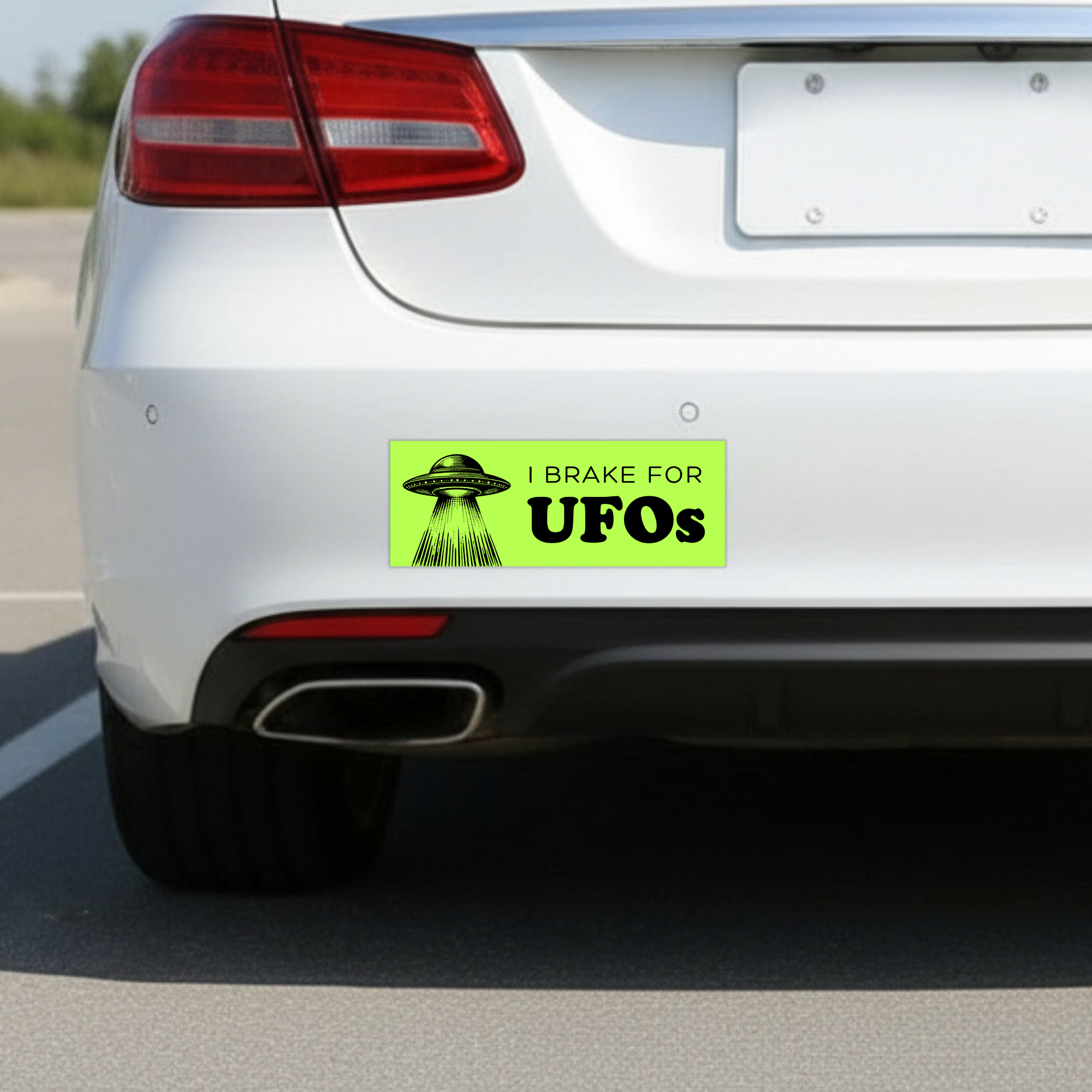 Sleazy Greetings funny bumper magnet displayed on the back of a white car featuring the phrase ‘I Brake For UFOs’ in bold black typography on a neon green background with a vintage-style flying saucer illustration beaming light downward. Humorous novelty car magnet with retro sci-fi aesthetic and alien conspiracy humor shown in real-life automotive use.
