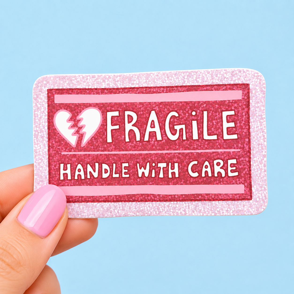 Sleazy Greetings funny holographic vinyl sticker styled like a shipping label featuring the phrase ‘Fragile Handle With Care’ with a broken heart icon. Glittery pink aesthetic sticker with emotional humor, soft pastel colors, and playful retro packaging design.
