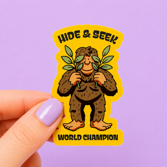 Sleazy Greetings funny die-cut vinyl sticker featuring a shy cartoon Bigfoot hiding behind leaves beside the phrase ‘Hide & Seek World Champion.’ Cute cryptid humor sticker with playful woodland artwork, goofy Sasquatch vibes, and retro outdoorsy aesthetic.