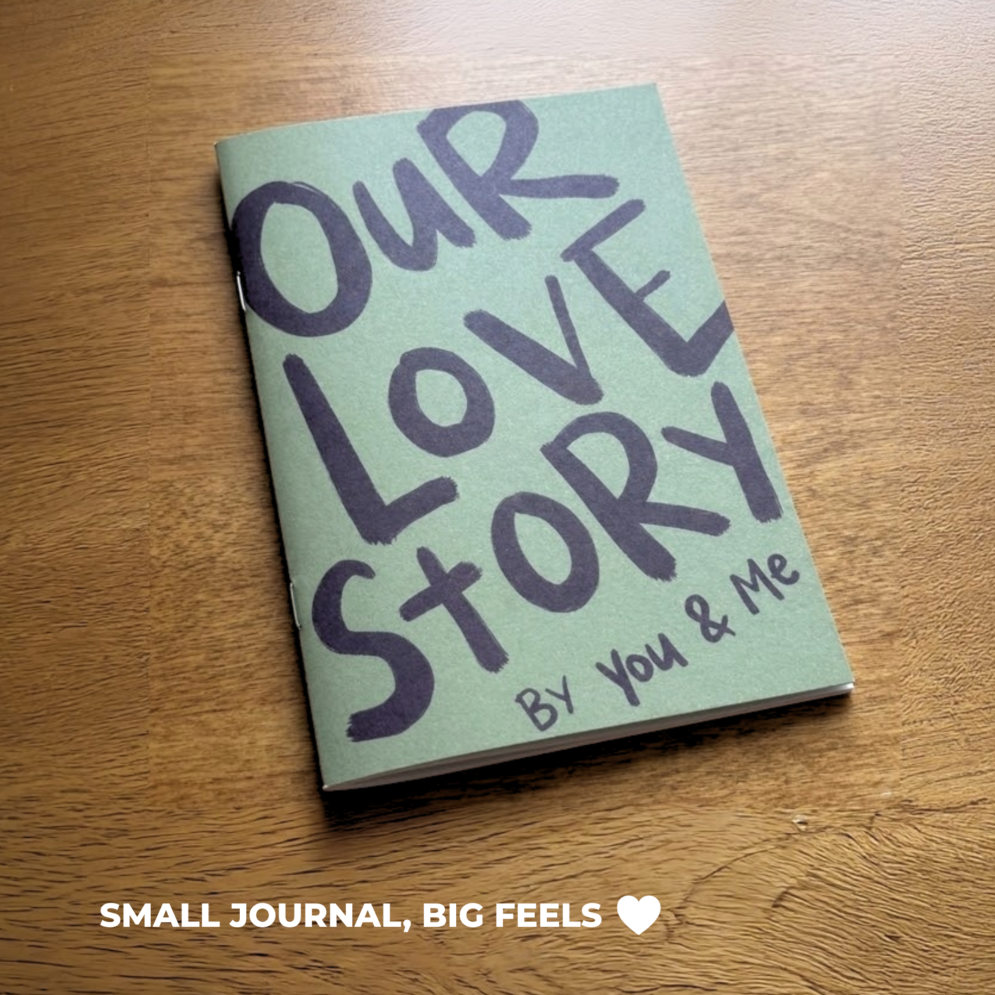 Sleazy Greetings ‘Our Love Story By You & Me’ compact couples journal resting on a wooden table, featuring bold hand-lettered dark purple typography on a muted green softcover. Small relationship memory book designed for couples to document shared stories, milestones, and meaningful experiences together.