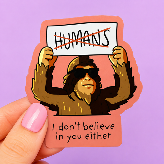 Sleazy Greetings funny die-cut vinyl sticker featuring a cool Bigfoot wearing sunglasses and holding a crossed-out ‘Humans’ protest sign beside the phrase ‘I Don’t Believe in You Either.’ Sarcastic cryptid humor sticker with playful cartoon artwork and rebellious paranormal vibes.
