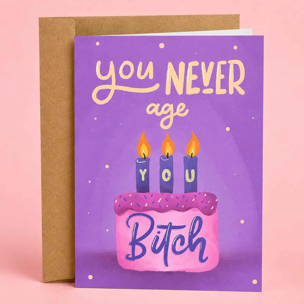 Sleazy Greetings funny birthday greeting card featuring a pink birthday cake with three lit candles spelling “YOU” and the sarcastic message “You Never Age, You Bitch” on a purple background, displayed with a kraft envelope for a humorous birthday card, gag gift, or funny birthday card for best friends, sisters, coworkers, and loved ones.