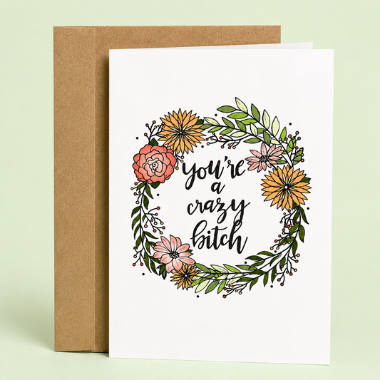 Sleazy Greetings funny friendship greeting card featuring a colorful floral wreath with hand-lettered typography reading “You’re a Crazy Bitch” on a white background, displayed with a kraft envelope for a humorous birthday card, best friend card, encouragement card, Galentine’s Day gift, or funny greeting card for someone who embraces their wild side.