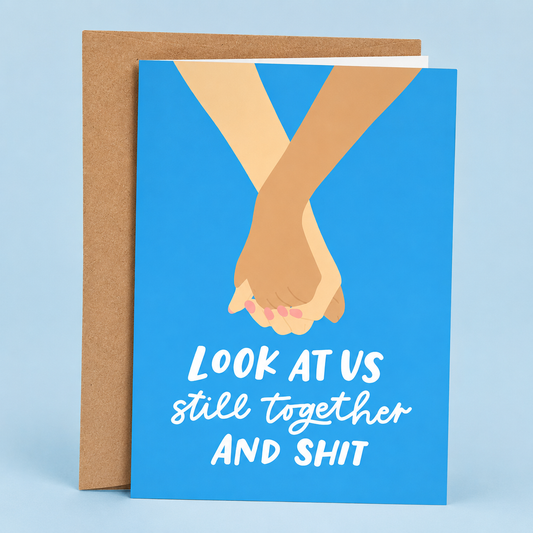 Sleazy Greetings funny love greeting card featuring two people holding hands on a bright blue background with the sarcastic romantic message “Look at Us Still Together and Shit,” displayed with a kraft envelope for an anniversary, Valentine’s Day, wedding, boyfriend, girlfriend, husband, or wife card.