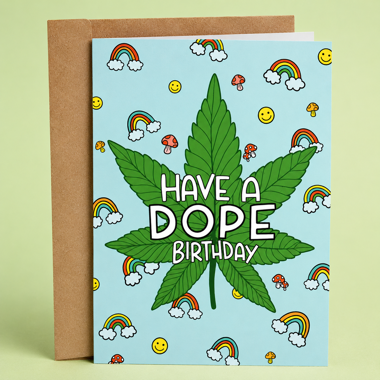 Sleazy Greetings funny cannabis birthday card featuring a large marijuana leaf on a light blue background surrounded by rainbows, smiley faces, and mushrooms. The card reads, “Have a Dope Birthday,” and is displayed with a kraft envelope.
