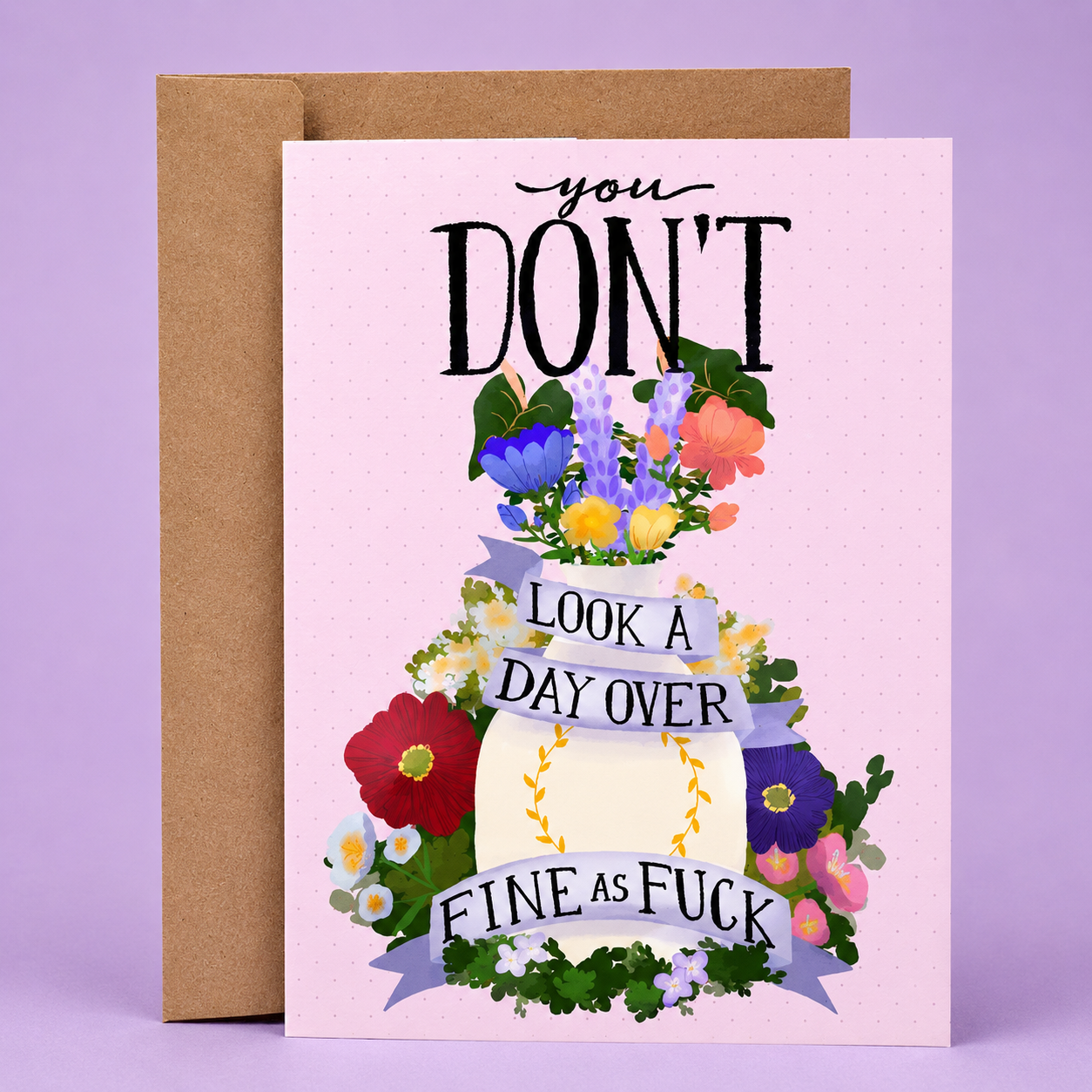 Sleazy Greetings funny encouragement card featuring a decorative floral arrangement with colorful flowers and ribbon banners on a pink background, with the message “You Don’t Look A Day Over Fine As Fuck,” paired with a kraft envelope.