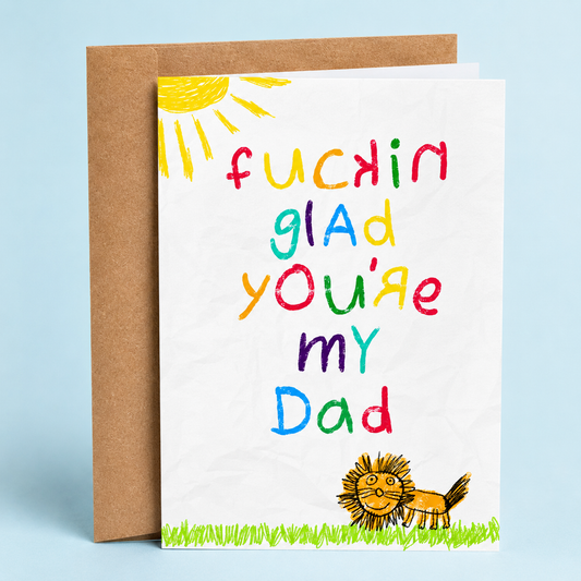 Sleazy Greetings funny Father’s Day greeting card featuring colorful childlike crayon lettering that reads “Fuckin Glad You’re My Dad” with a doodle-style lion, sunshine, and grass drawing on a white crumpled paper background, paired with a kraft envelope.