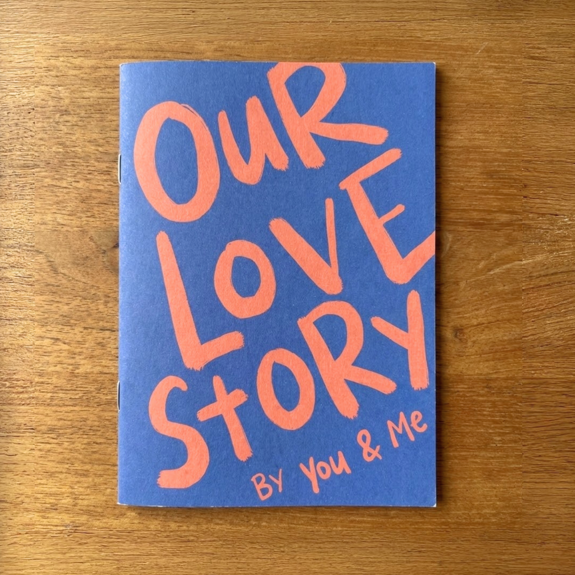 Sleazy Greetings ‘Our Love Story By You & Me’ couples journal featuring bold coral hand-lettered typography on a navy blue softcover, displayed on a wooden surface. Relationship memory book designed for couples to document shared adventures, milestones, and meaningful experiences together.