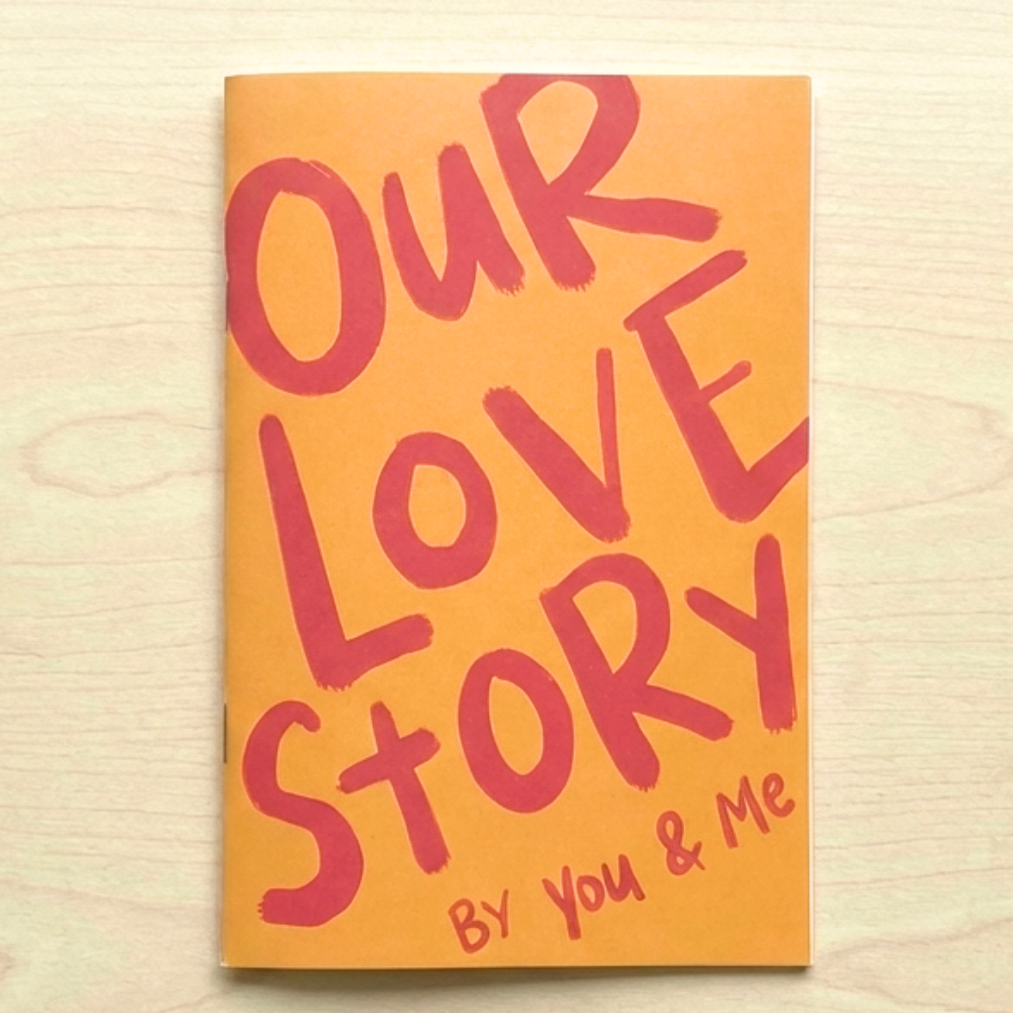 Sleazy Greetings ‘Our Love Story By You & Me’ couples journal featuring bold hand-lettered pink typography on a vibrant orange softcover, displayed on a light wood surface. Fun relationship keepsake journal designed for couples to record memories, adventures, milestones, and meaningful shared experiences together.